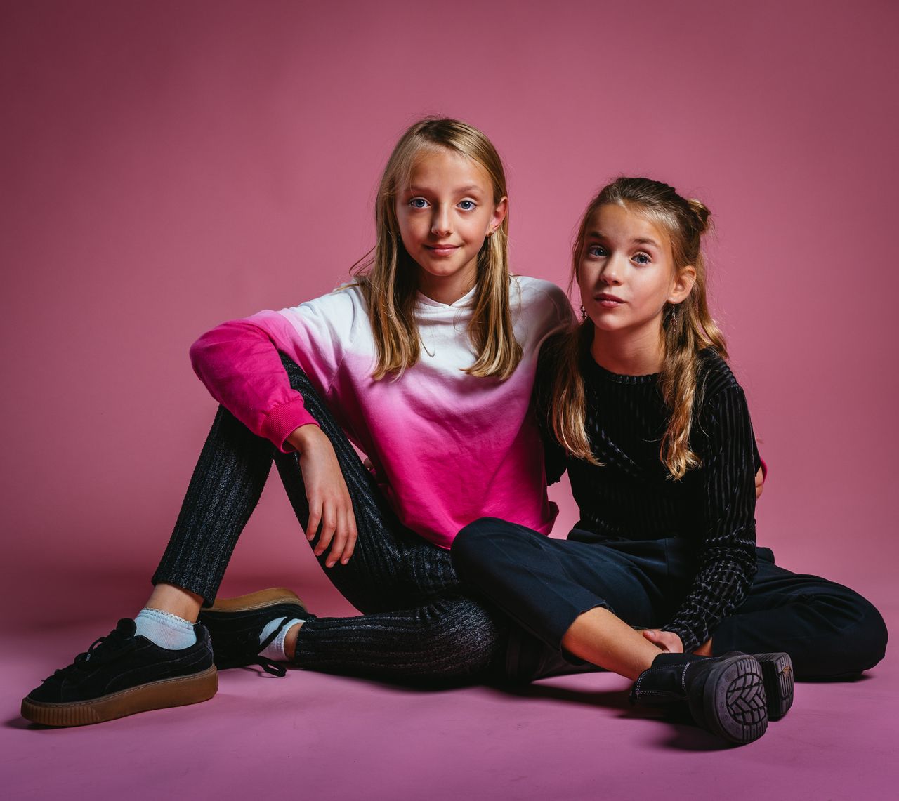 Charming portrait of two young girls sitting in a studio with a pink backdrop, showcasing casual and stylish clothing.