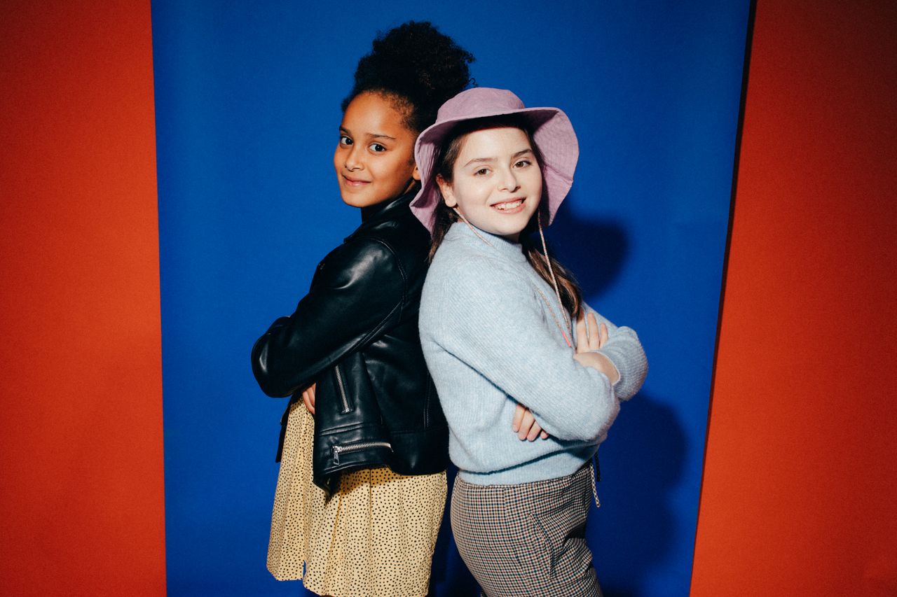 Two girls posing back-to-back in fashionable outfits against a colorful backdrop.
