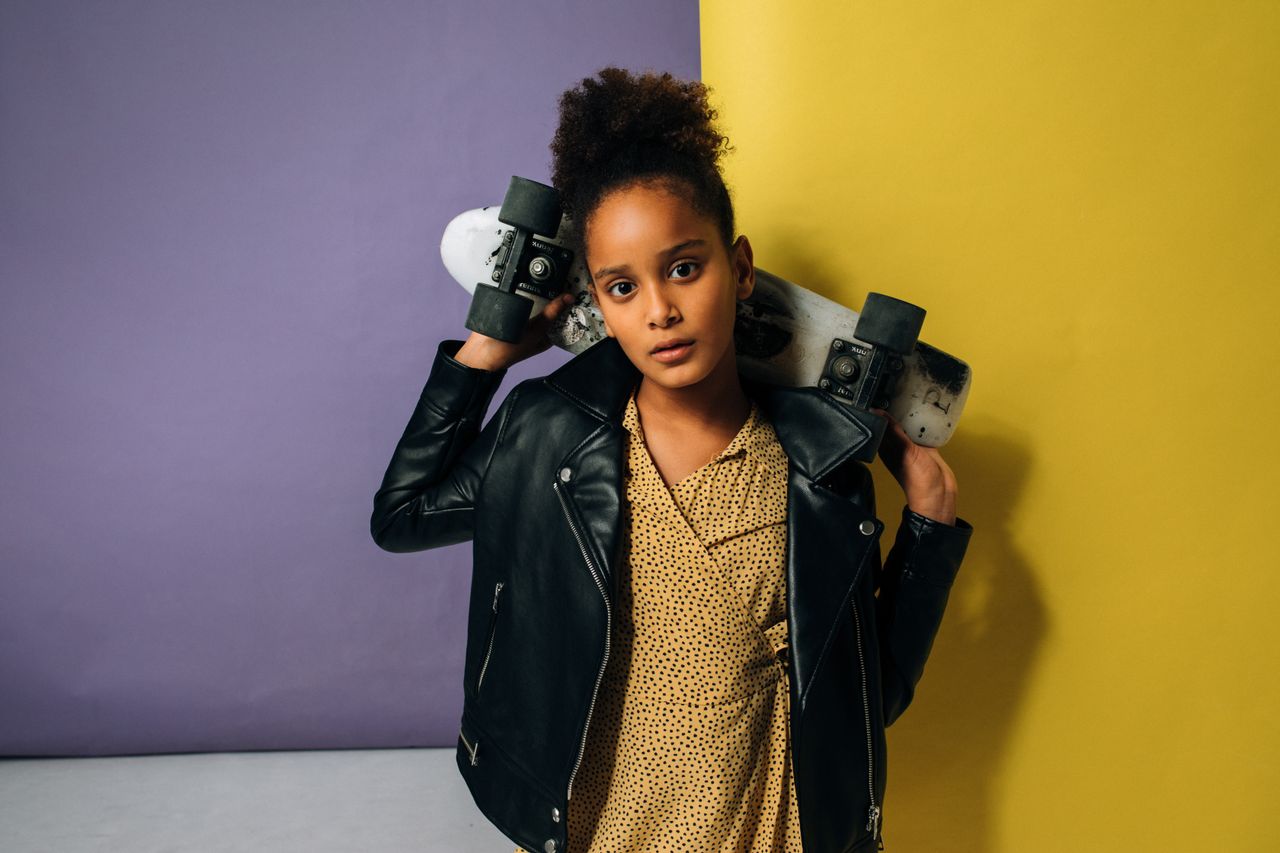 Confident young girl with skateboard and leather jacket posing indoors.